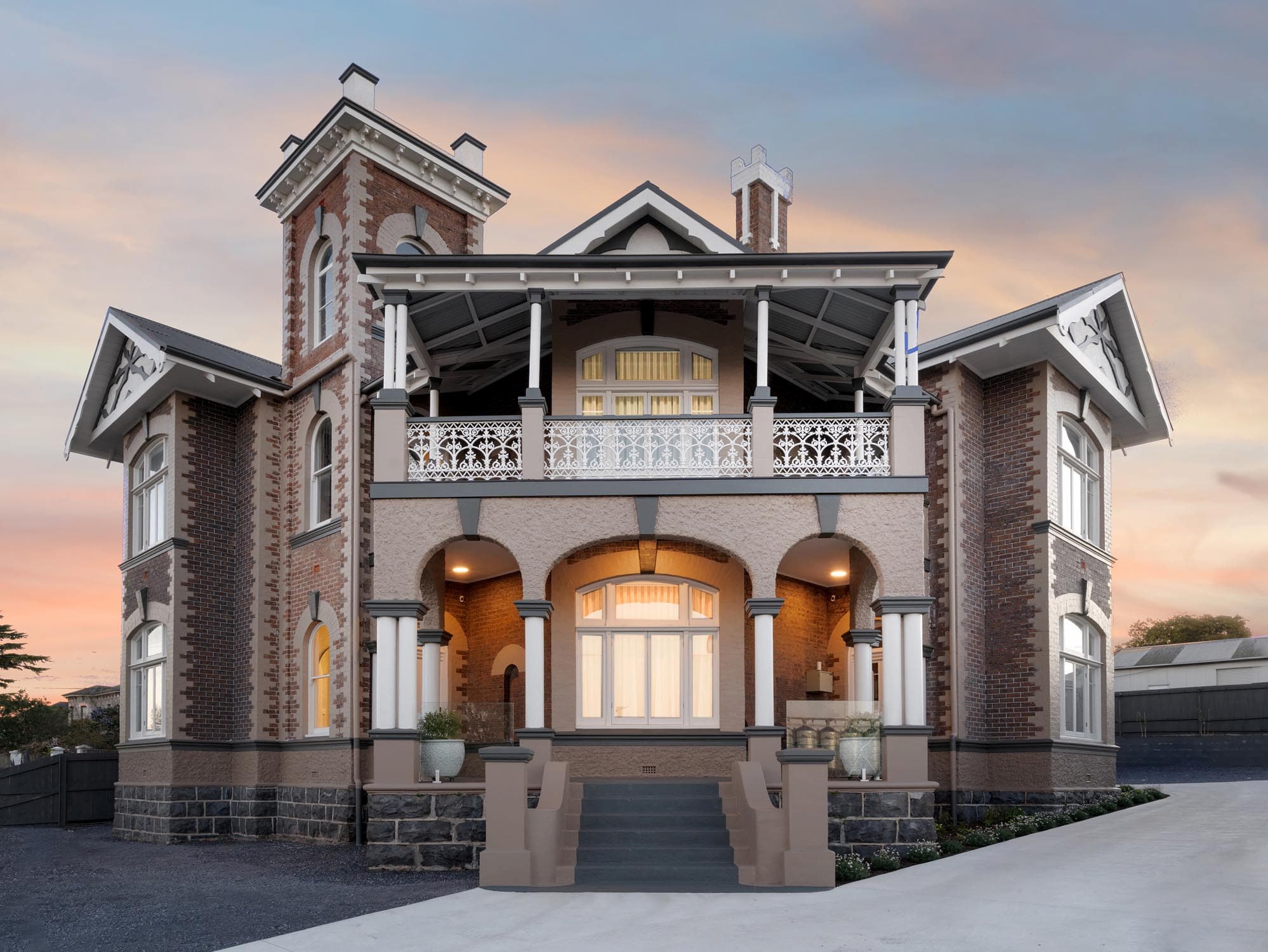 A large, historic two-storey brick house features a central tower, arched entryway, upper balcony with decorative railing, and detailed trim, all set against a sunset sky.