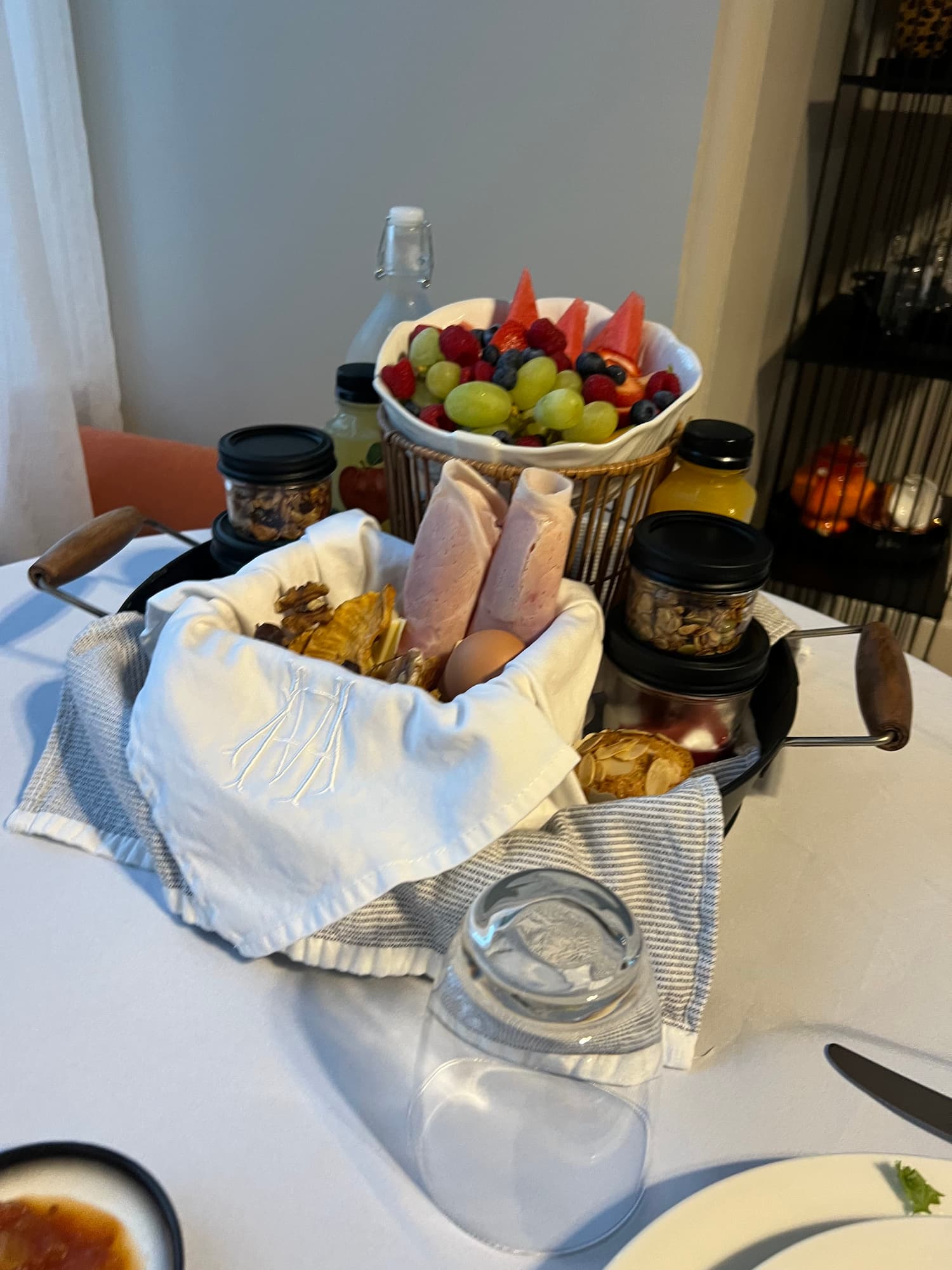 A breakfast spread with assorted cold meats, hard-boiled eggs, bread, fresh fruit, small jars, and bottles of juice arranged on a white tablecloth.