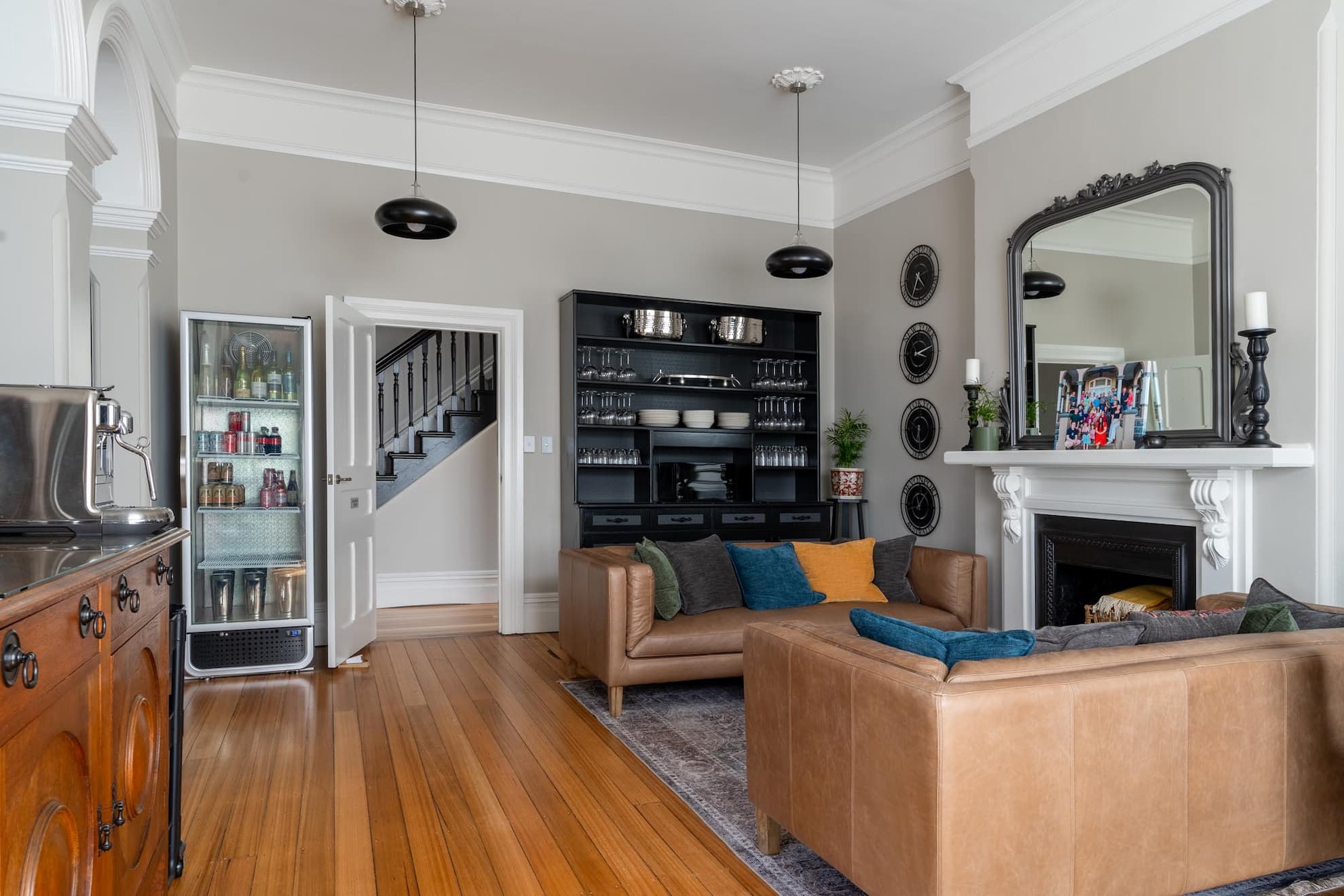 A living room with tan leather sofas, a dark display cabinet with crockery, a fireplace with a mirror above, a small fridge with drinks, and wooden floors. The room has neutral walls and two black pendant lights.
