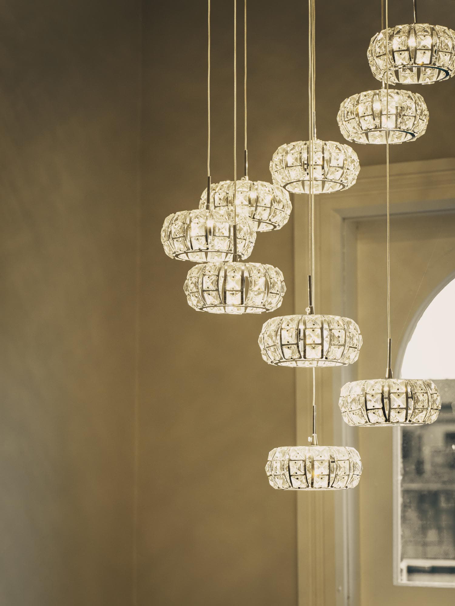 Several round crystal pendant lights hang at varying heights in front of a pale wall and a window with an arched top, creating a modern and elegant lighting arrangement.