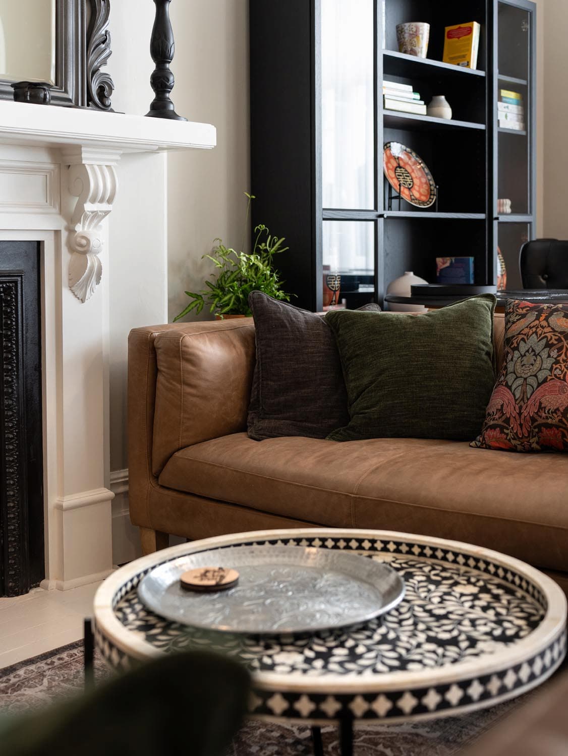 A tan leather sofa with assorted cushions sits beside a round patterned coffee table holding a metal tray and a small dish. A black shelving unit with books and decor stands against the wall near a white fireplace.
