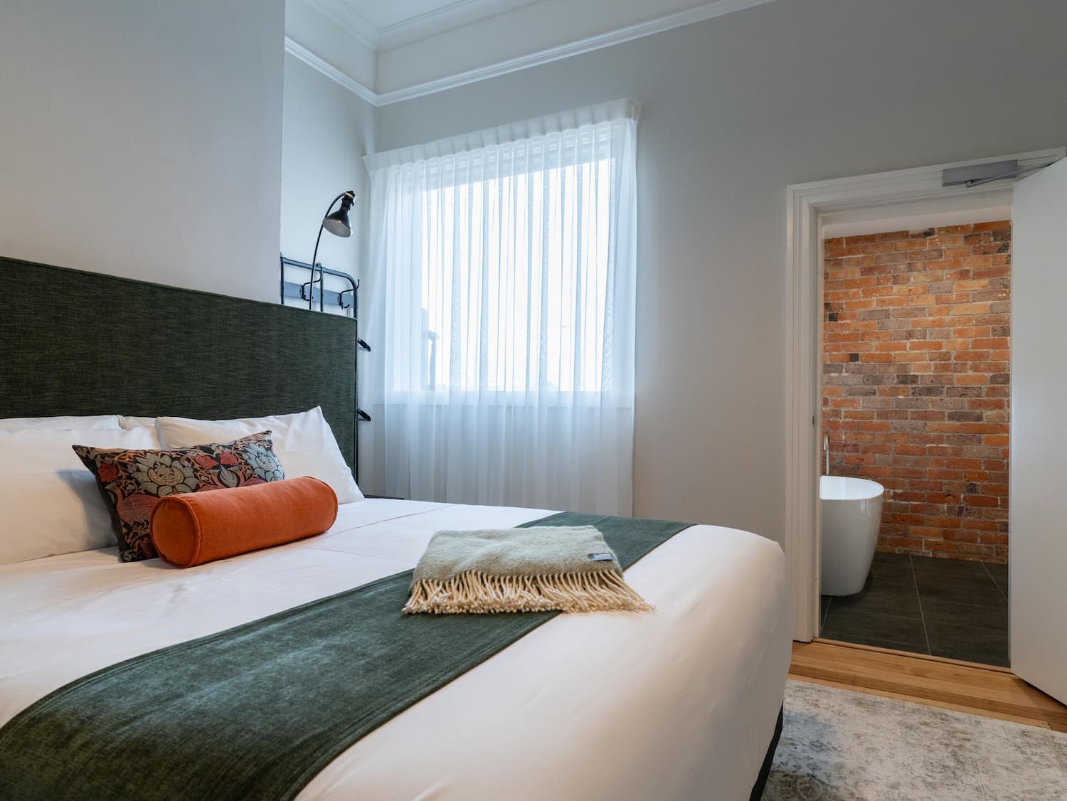 A modern bedroom with a large bed, green accents, and a throw blanket; an open door reveals a bathroom with exposed brick walls and a freestanding bath.