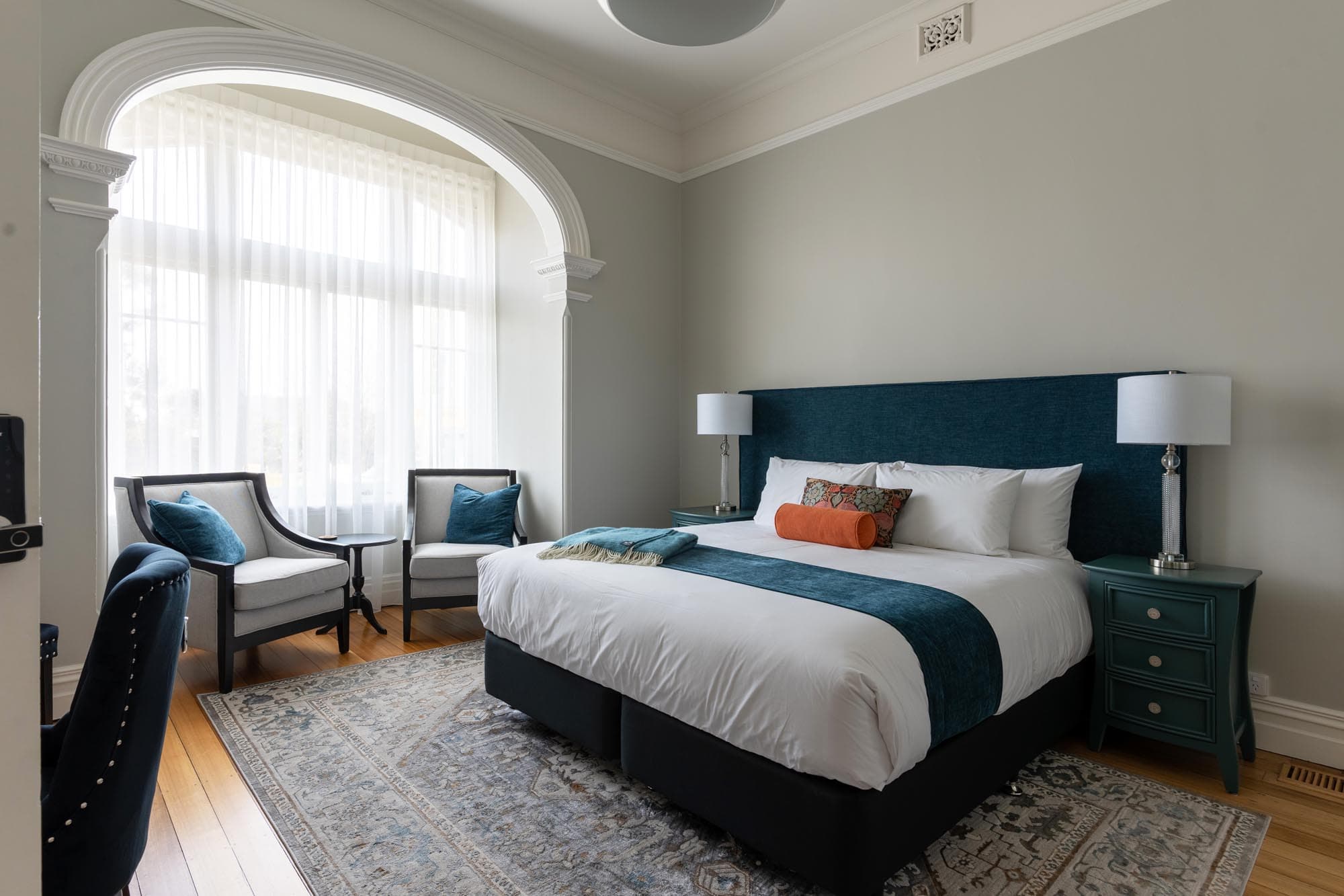 A bedroom with a large bed, teal upholstered headboard, teal bedside table, two armchairs by a bright arched window with sheer curtains, and a patterned rug on a wooden floor.
