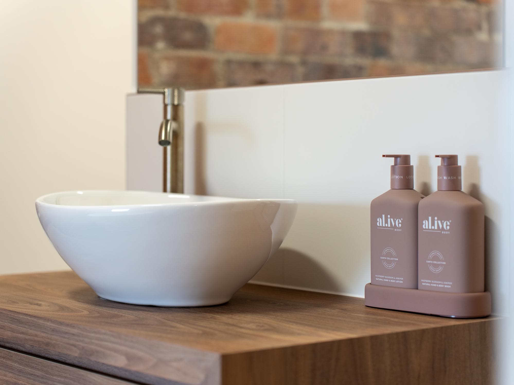 A modern bathroom washbasin with a round white basin on a wooden worktop, a brass tap, and two mauve-coloured soap dispensers beside the basin.