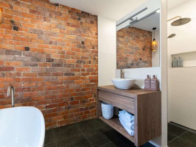 Modern bathroom with exposed brick wall, vessel basin on a wooden vanity unit, stacked towels on an open shelf, white bath, and a large mirror above the basin. A glass shower with a rain showerhead is visible in the background.